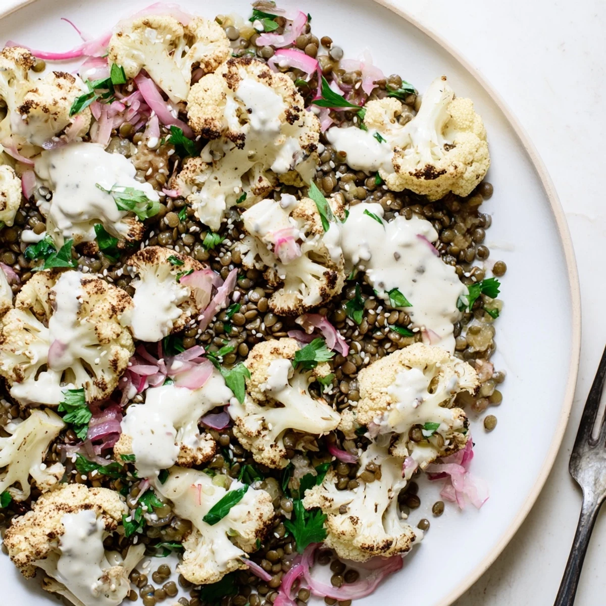 A close-up view of a vibrant vegetarian bowl featuring spiced roasted cauliflower, zesty lemon lentils, and a tangy cumin tahini yogurt drizzle.