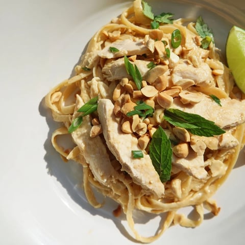 Steaming bowl of Thai Peanut Chicken Pasta, aromatic herbs, peanuts, and lime wedges.