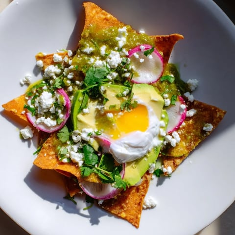Crispy tortilla chips coated in zesty salsa verde, topped with a runny fried egg, avocado slices, and crumbled queso fresco on a plate.  