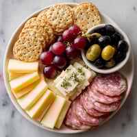 A plate of food with cheese, crackers, grapes, olives, and meat.